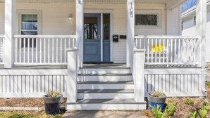A white two-story house with a front porch, a blue door, and a small garden with potted plants.