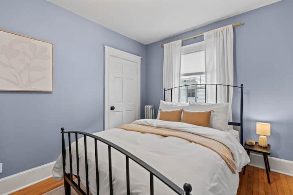 A bedroom with a bed, a nightstand, a window with curtains, a door, and a wall-mounted artwork.