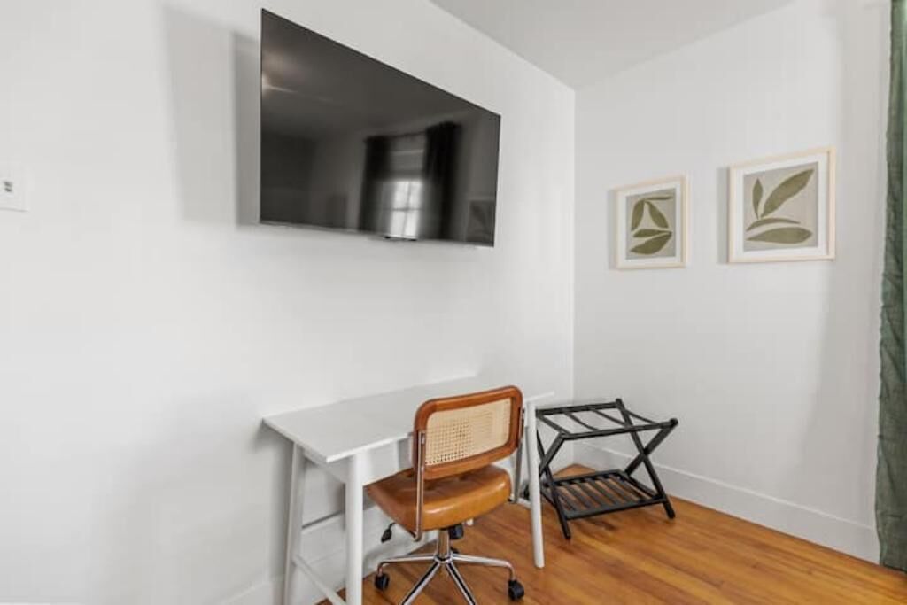 Ein Zimmer mit einem weißen Schreibtisch, einem Korbstuhl, einem schwarzen Metallregal und zwei gerahmten Blattform-Motiven an der Wand.
