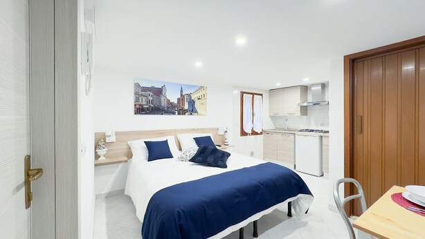 A hotel room with a bed, a small table, a chair, a kitchenette, and a door.