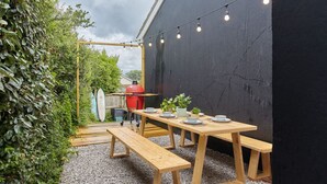Ein Essbereich im Freien mit Holzbänken und einem Tisch für zwei Personen, umgeben von Grünpflanzen und einer schwarzen Wand.
