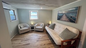 A cozy room with a wooden ceiling, a large painting of a pier, a white sofa, two armchairs, and a small table with a lamp. - Adorable beach house - 5 min walk to private beach (Wareham)