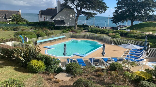 En swimmingpool med blå og hvide liggestole, omgivet af et hvidt hegn, med udsigt til havet og huse i baggrunden.