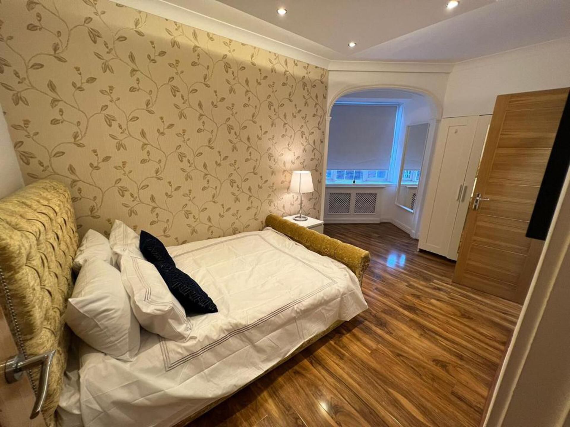 A bedroom with a bed, a nightstand, a lamp, and a view of a balcony through an archway.