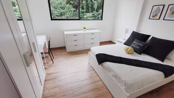 A modern bedroom with a large bed, a white dresser, a small desk, and a window with a view of greenery.