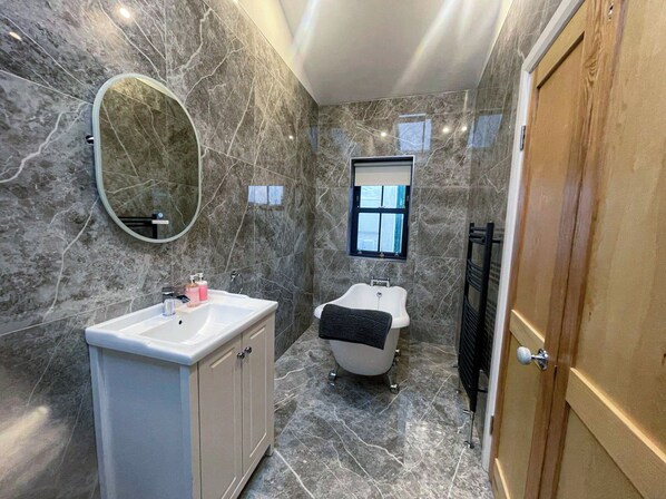 A bathroom with a marble floor, a white sink with a mirror, a bathtub, and a window. - Bayview Holiday Home (Dunmore East)