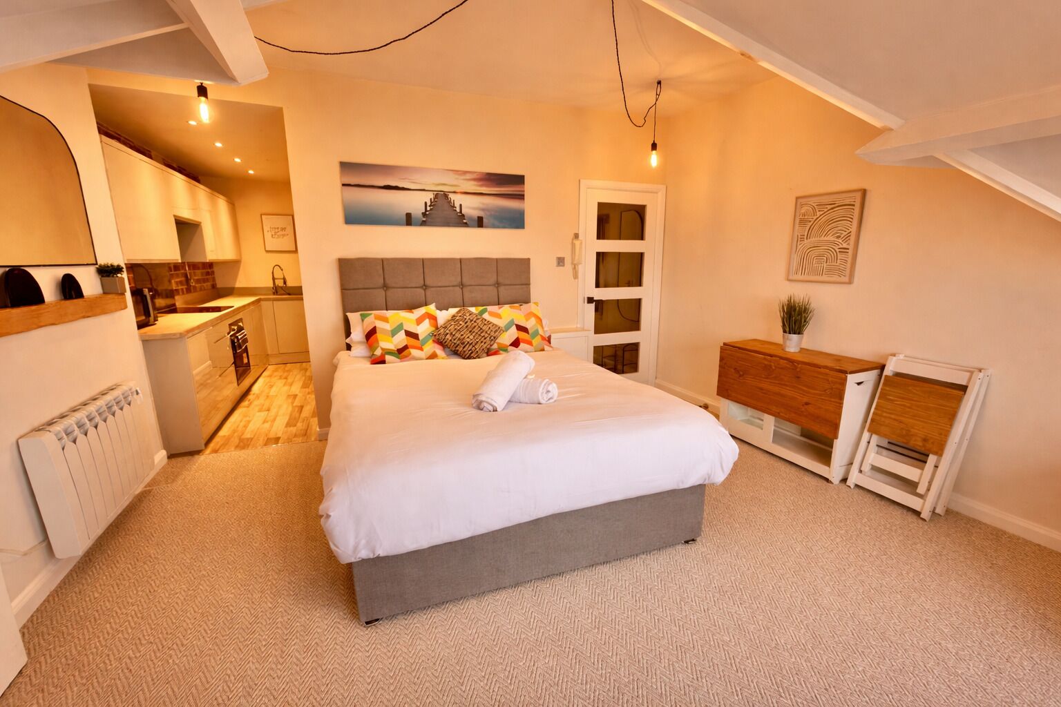 A bedroom with a bed, a bedside table, a dresser, a radiator, a kitchen area, and a door.