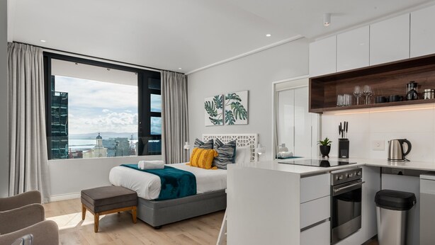 A modern hotel room with a large window, a bed with a view, a kitchenette, and a seating area. - Bright Studio With City Views - No Load-shedding (Cape Town)