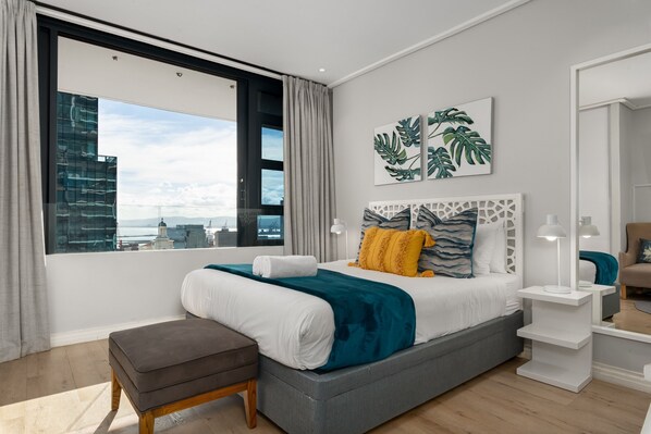 Camera da letto moderna con un'ampia finestra, un letto grigio, biancheria da letto bianco e azzurro e vista sulla città.