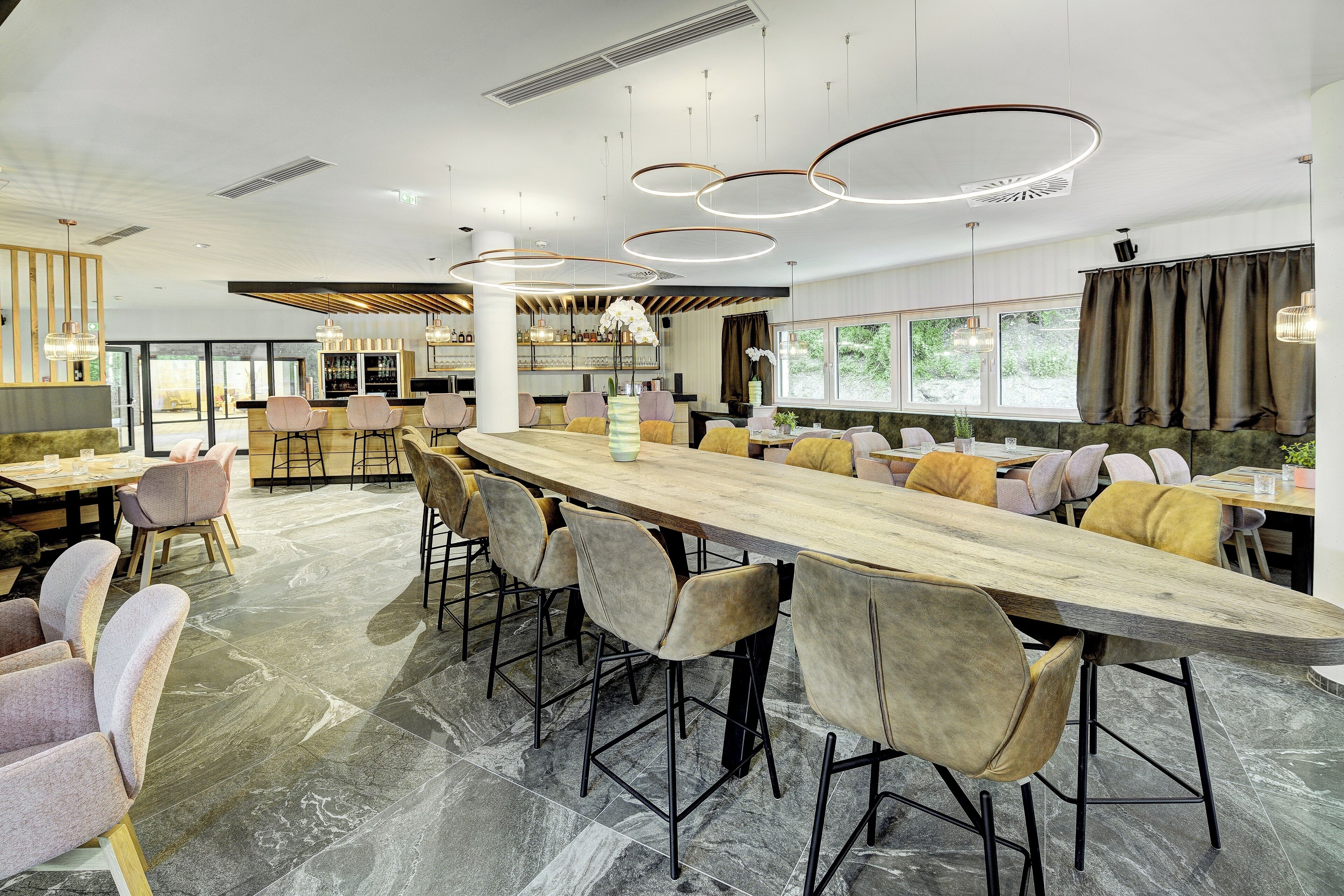 A modern restaurant with a long communal table, cushioned chairs, and a bar area.