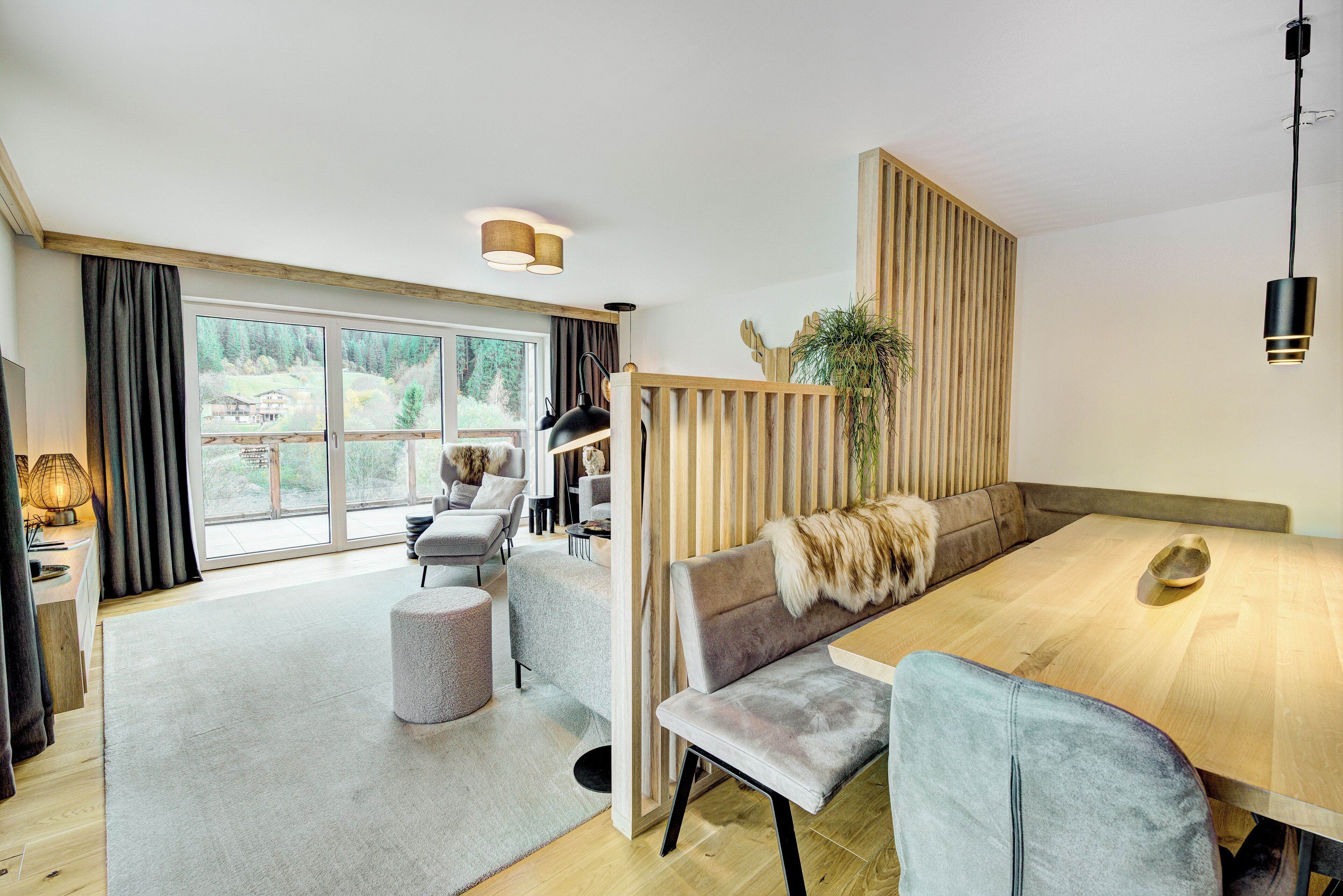 A modern living room with a wooden dining table, a grey sofa, and a large window offering a view of a forested area.