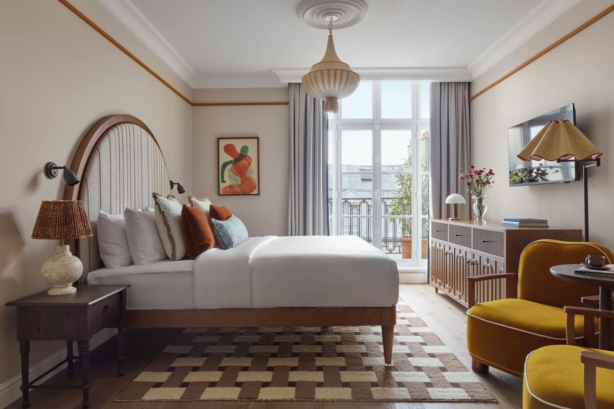 A modern bedroom with a large bed, a sofa, a yellow armchair, a small table, a lamp, a painting, a TV, and a view of the outside.