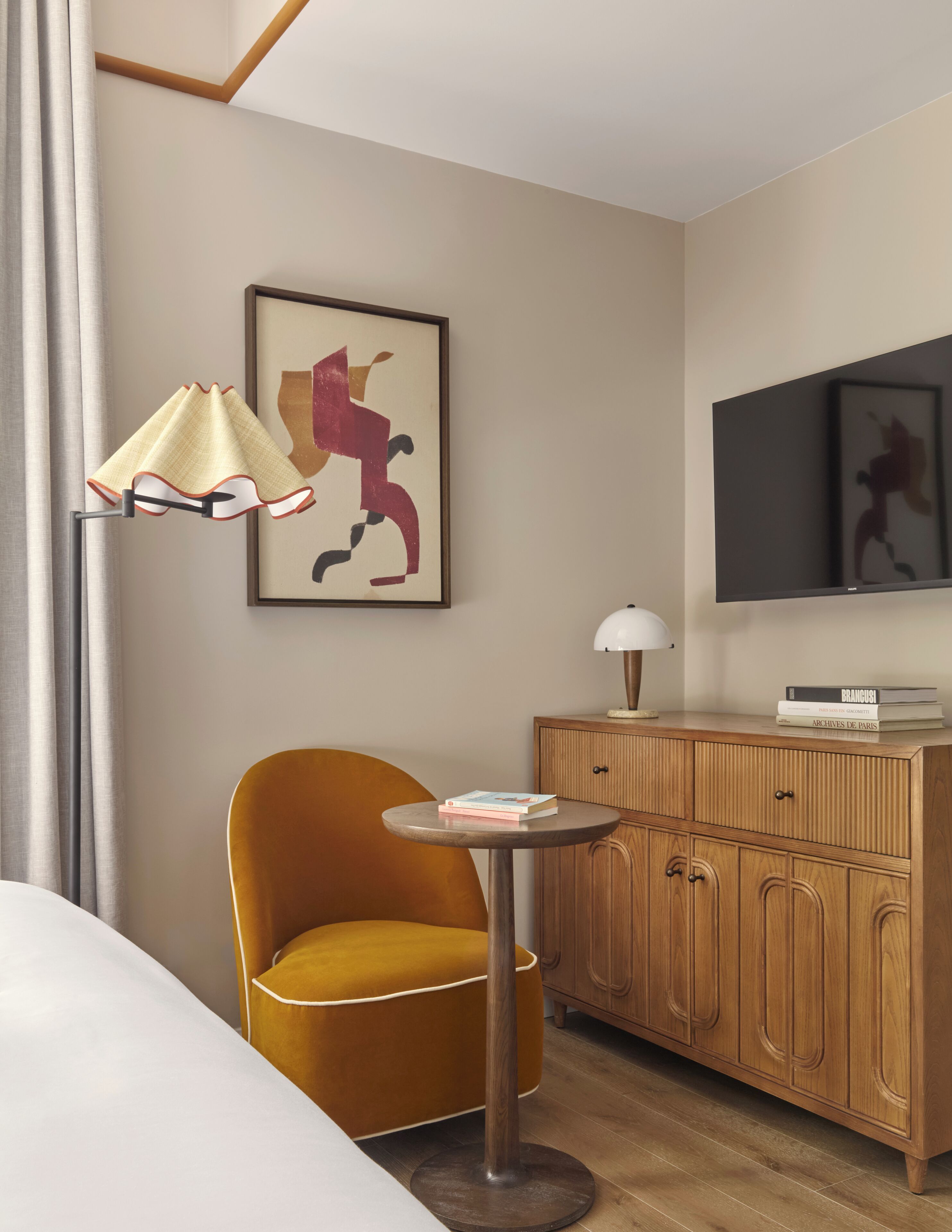 Een hotelkamer met een gele fauteuil, een klein rond tafeltje met boeken, een houten kast en een aan de muur bevestigde televisie.