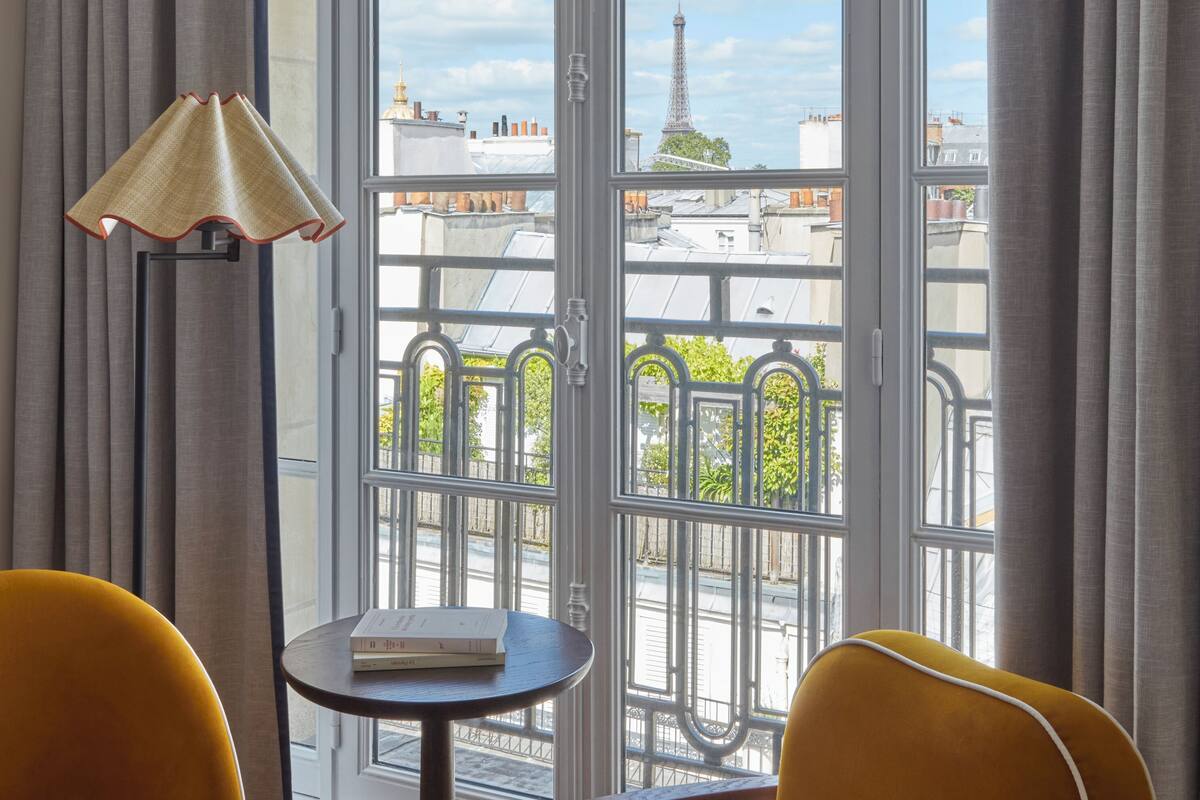 A room with two yellow chairs, a small table with a book, and a lamp. A large window with a view of the Eiffel Tower is visible.