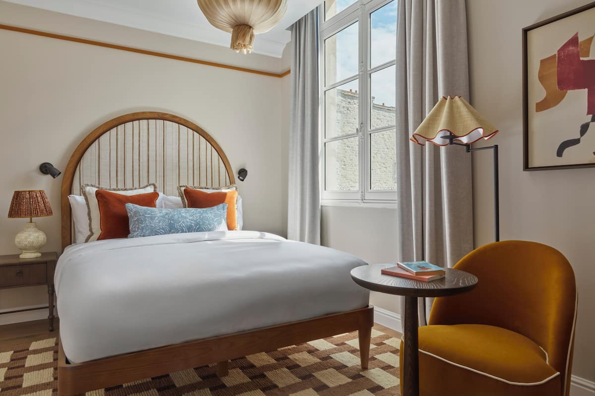 A hotel room with a large bed, a round table, a chair, a lamp, and a window with curtains.