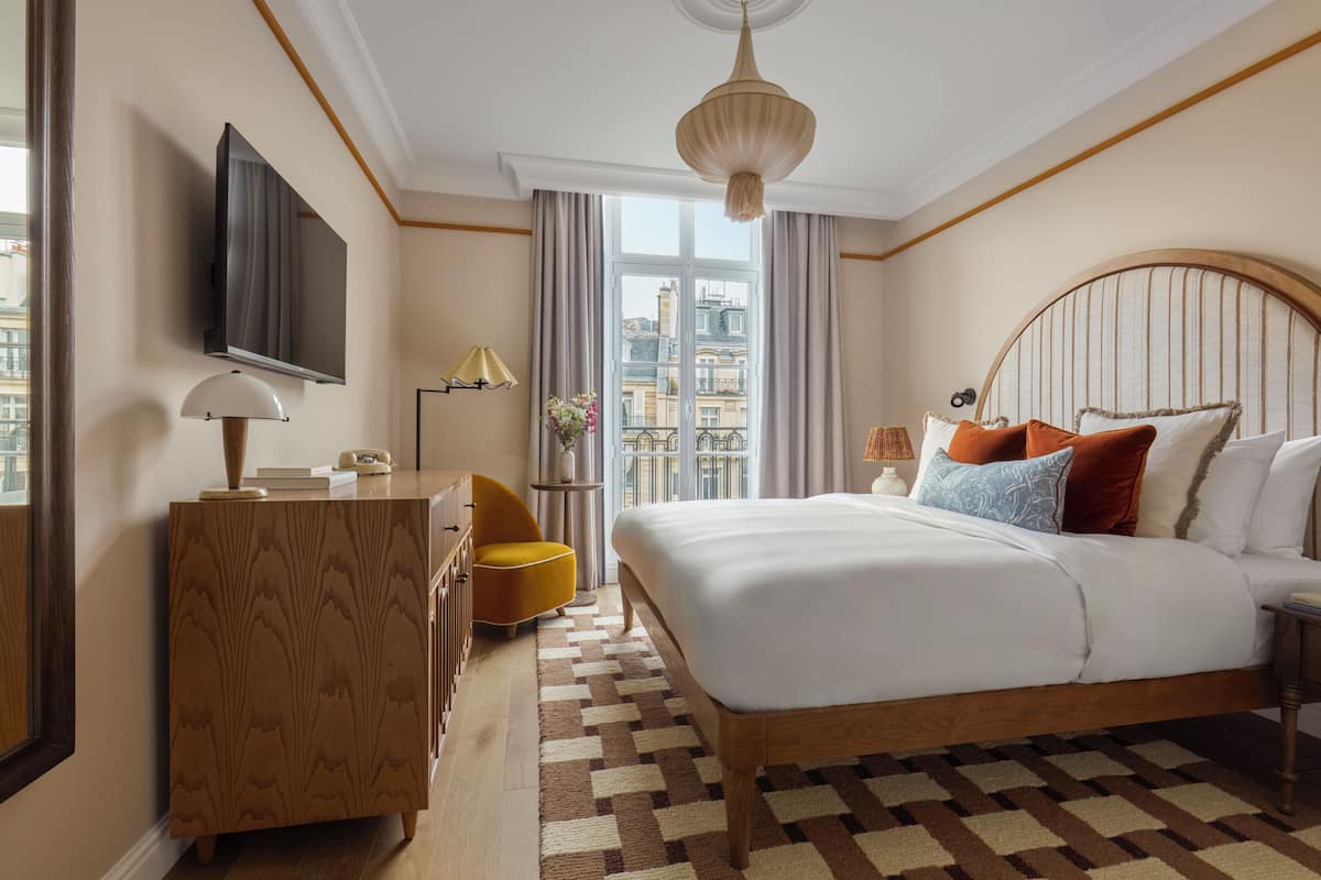 A hotel room with a large bed, a TV, a desk, a chair, and a view of the city through the window.