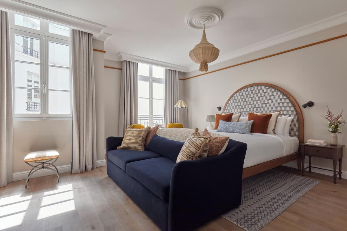 A bedroom with a large bed, a dark blue sofa, a small stool, and a bedside table with a lamp.