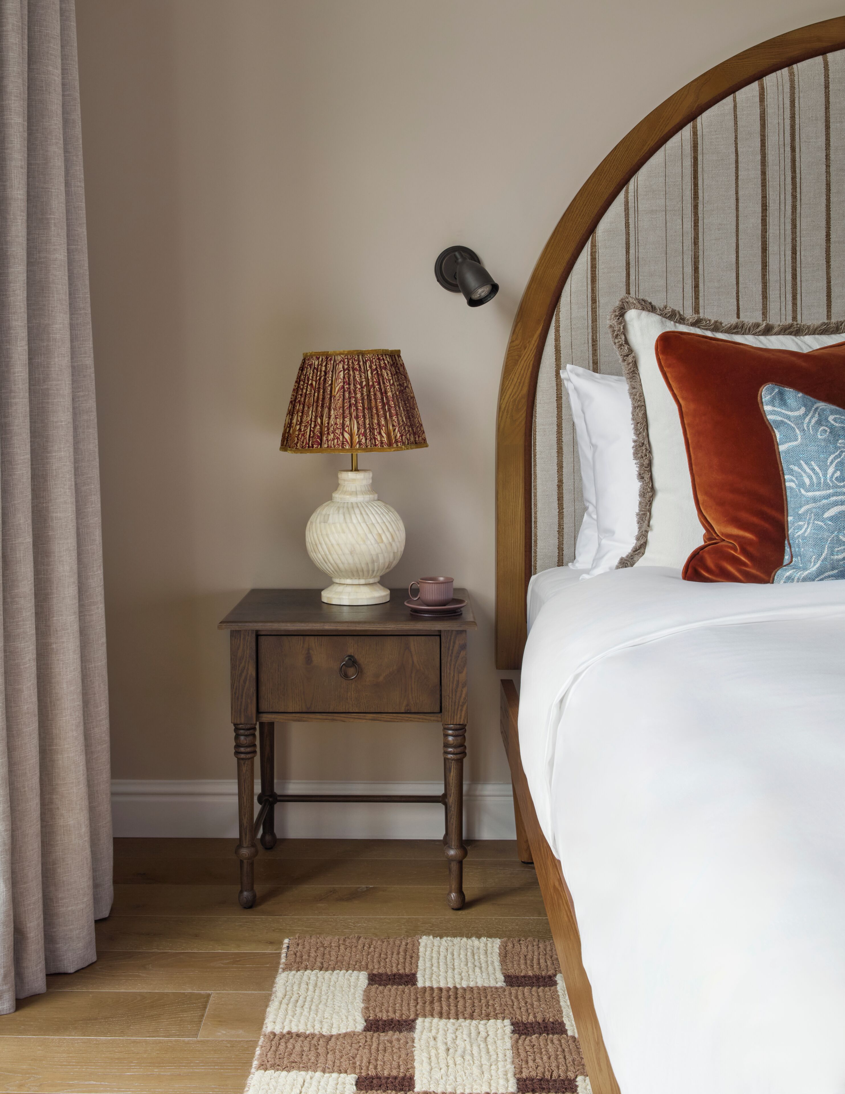 A bedroom with a wooden nightstand, a white lamp, a patterned rug, and a bed with a headboard.