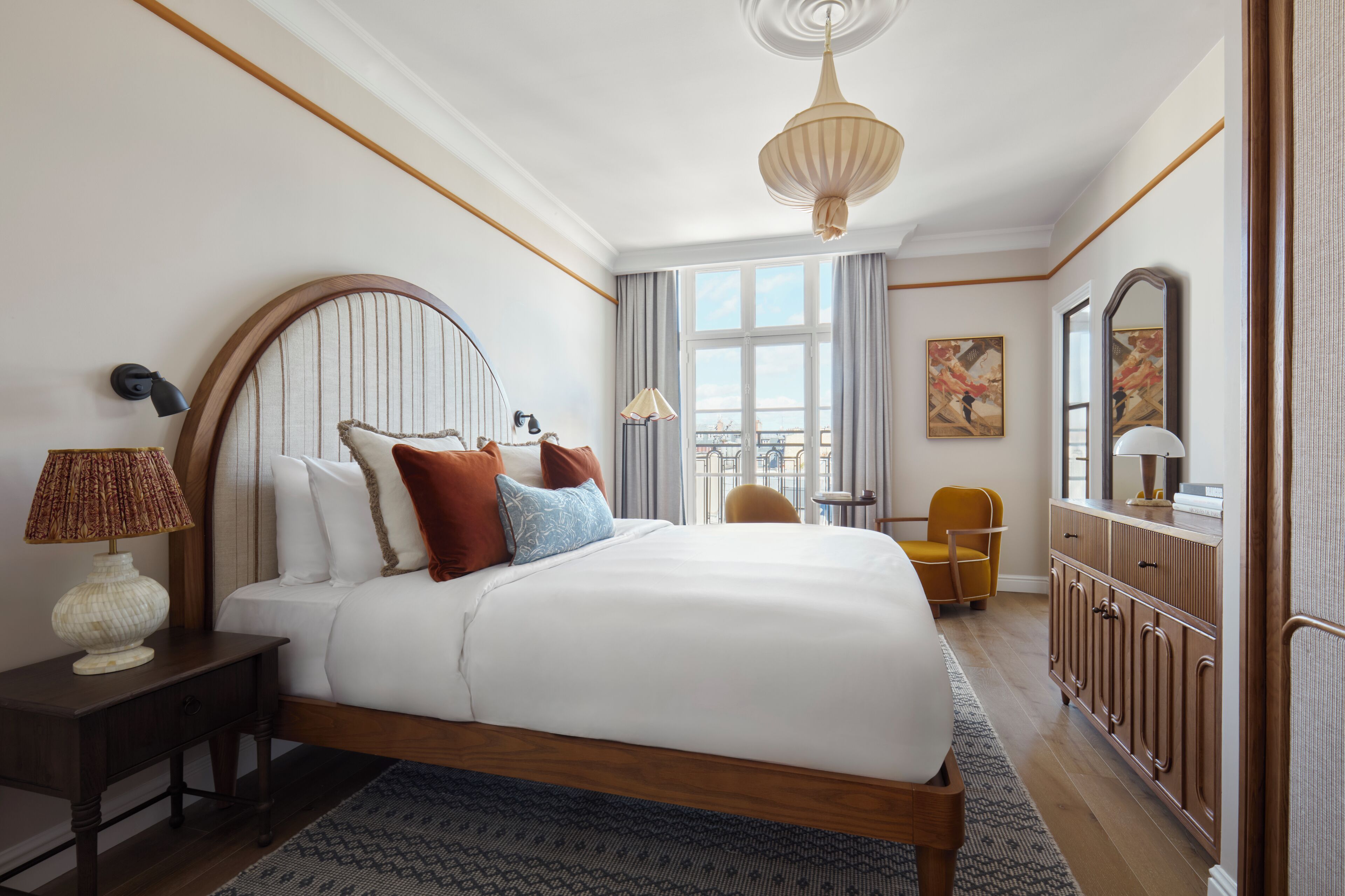A hotel room with a large bed, a wooden headboard, a nightstand, a lamp, a chair, a dresser, and a large window with curtains.