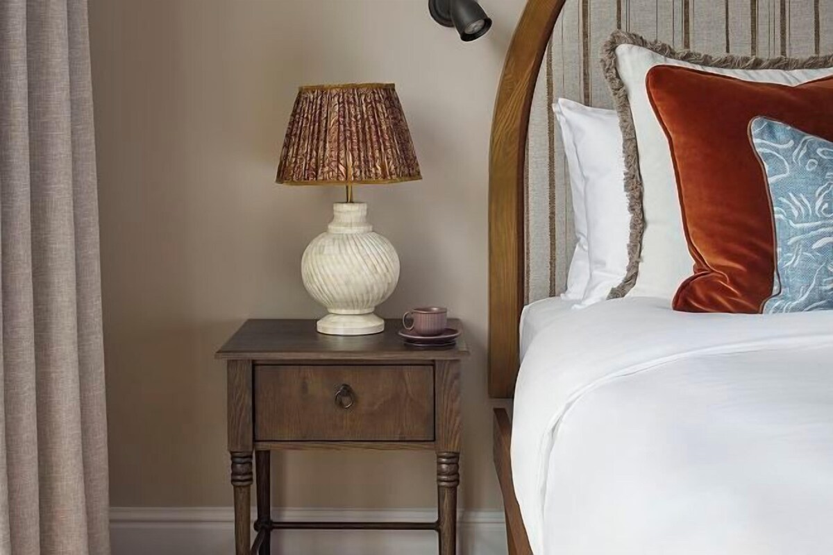 A neatly made bed with a white comforter and a brown headboard. A small bedside table with a lamp and a cup is visible.