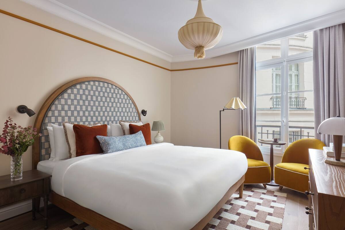 A hotel room with a large bed, two yellow chairs, a desk, a lamp, and a view of the city through large windows.