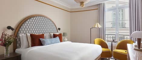A hotel room with a large bed, two yellow chairs, a desk, a lamp, and a view of the city through large windows.