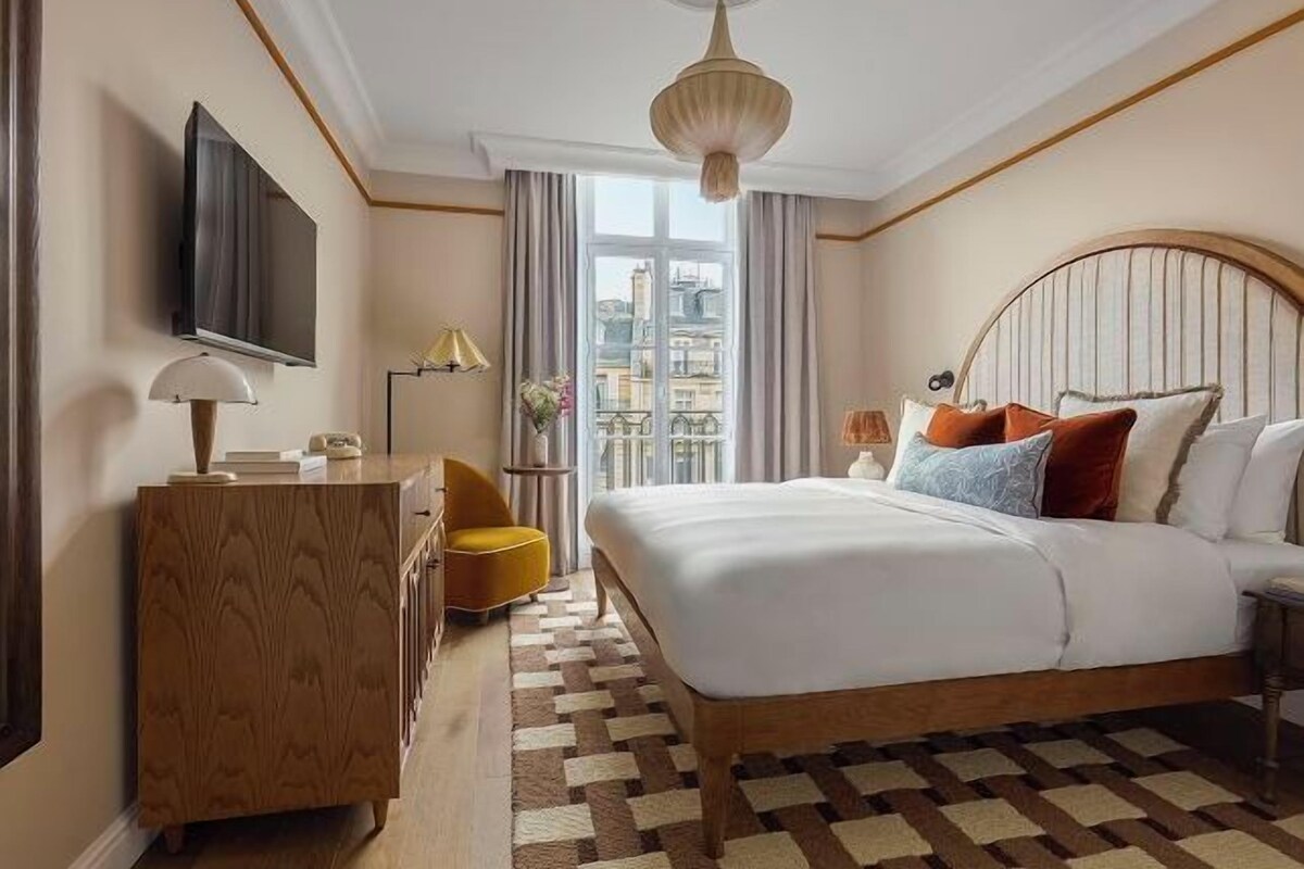 A hotel room with a large bed, a wooden dresser, a television, and a view of the city through the window.