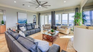 A spacious living room with a large gray sofa, a smaller sofa, and a coffee table. There is a ceiling fan, large windows with curtains, and a view of the ocean. - Oyster House B2 - Sleeps 20 - Beachfront w/ Pool (Gulf Shores)