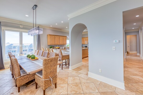 A spacious dining area with a long wooden table and wicker chairs, a chandelier, and a view of the outside through large windows. - Oyster House B2 - Sleeps 20 - Beachfront w/ Pool (Gulf Shores)