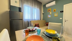 Un espace de vie compact comprenant un coin cuisine, un coin repas avec une table pour deux personnes, un canapé et un climatiseur fixé au mur.