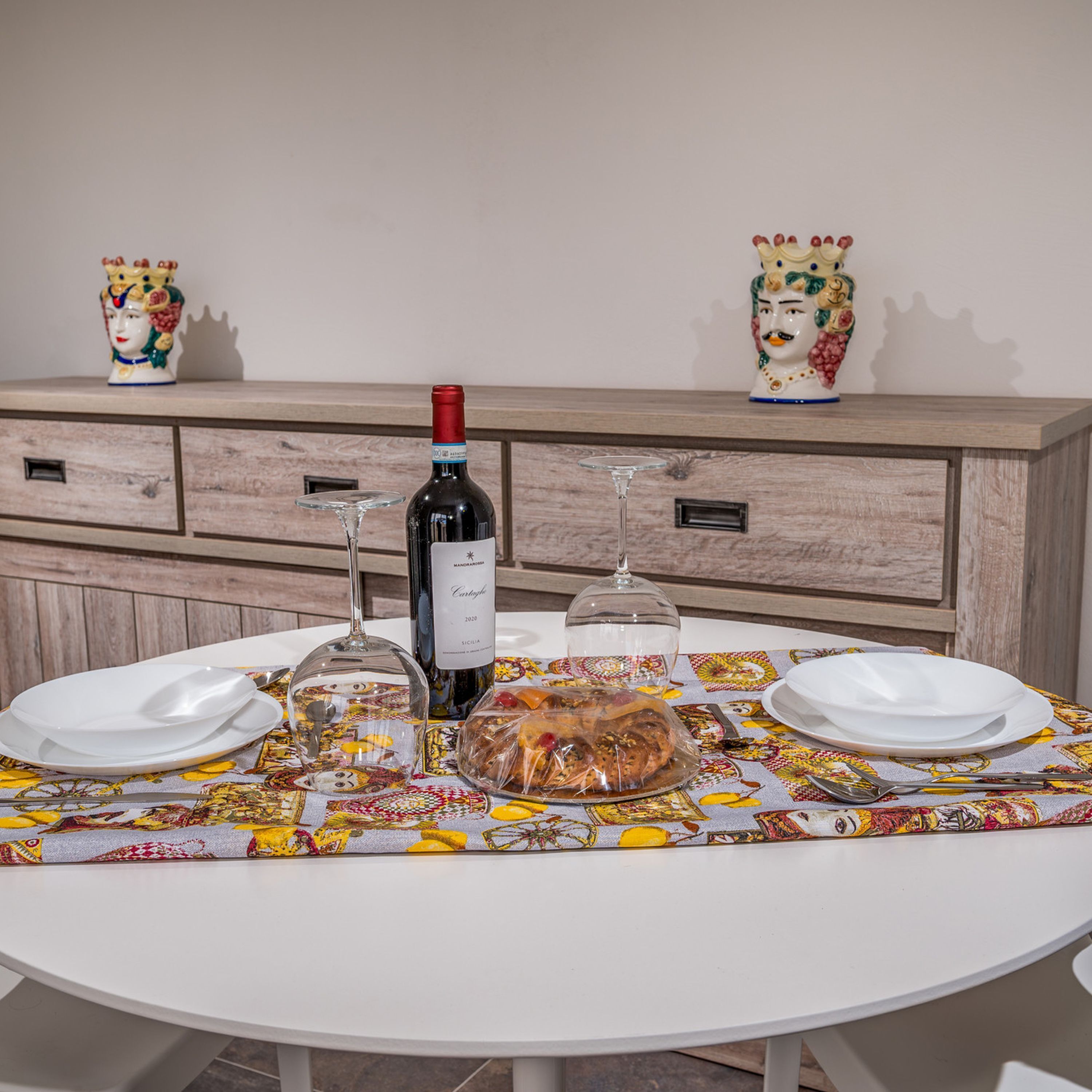 A round dining table with a floral tablecloth, two white plates, a bottle of wine, a glass, and a covered food item.