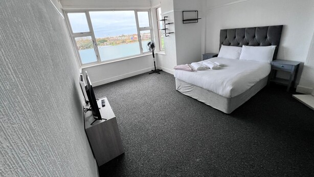 A modern hotel room with a large bed, a desk with a computer, a window with a view, and a fan. - Harbour Stays (Skegness)