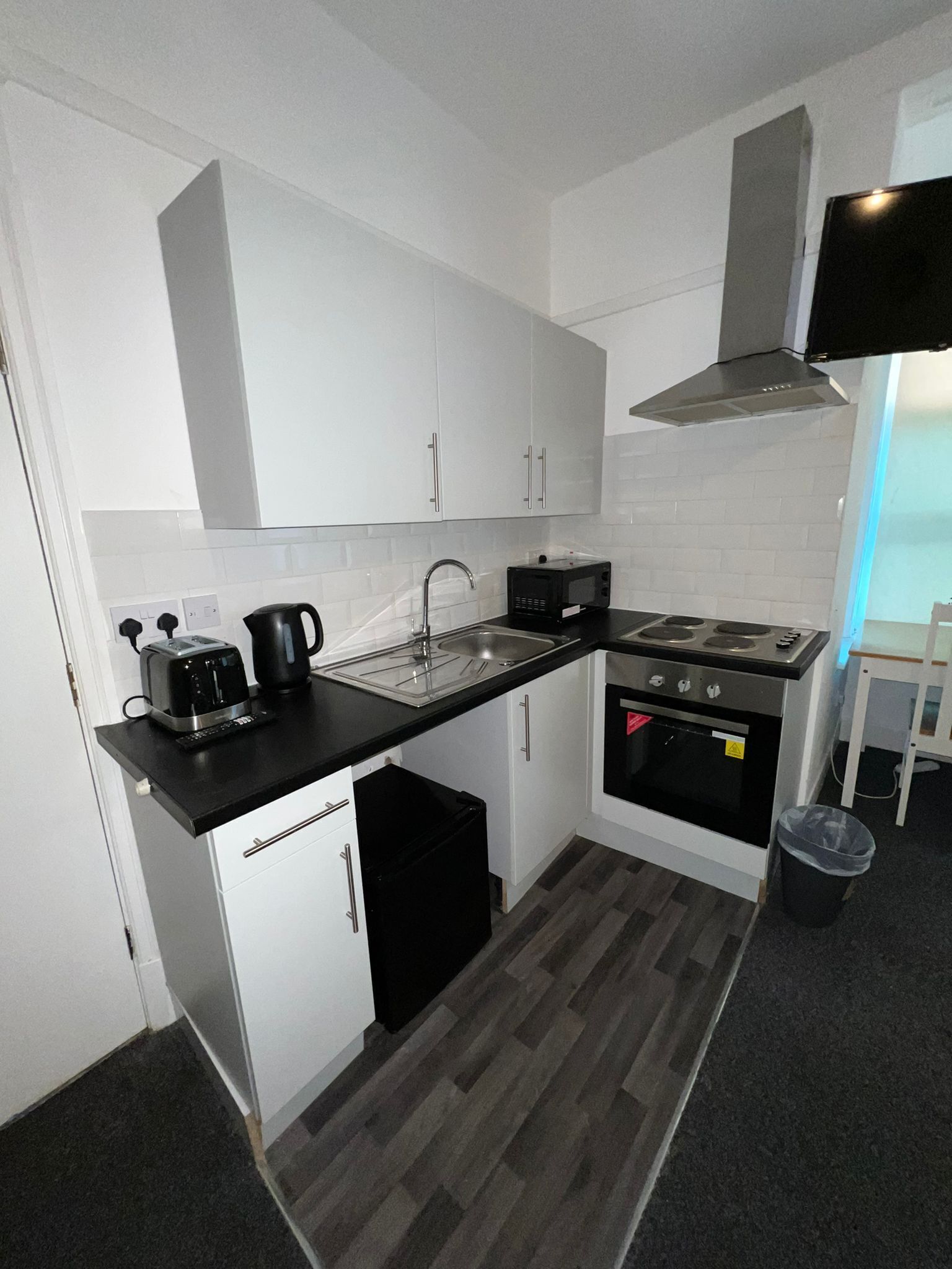 Une cuisine de taille réduite avec des meubles blancs, un plan de travail noir, une cuisinière, un four à micro-ondes et une poubelle.