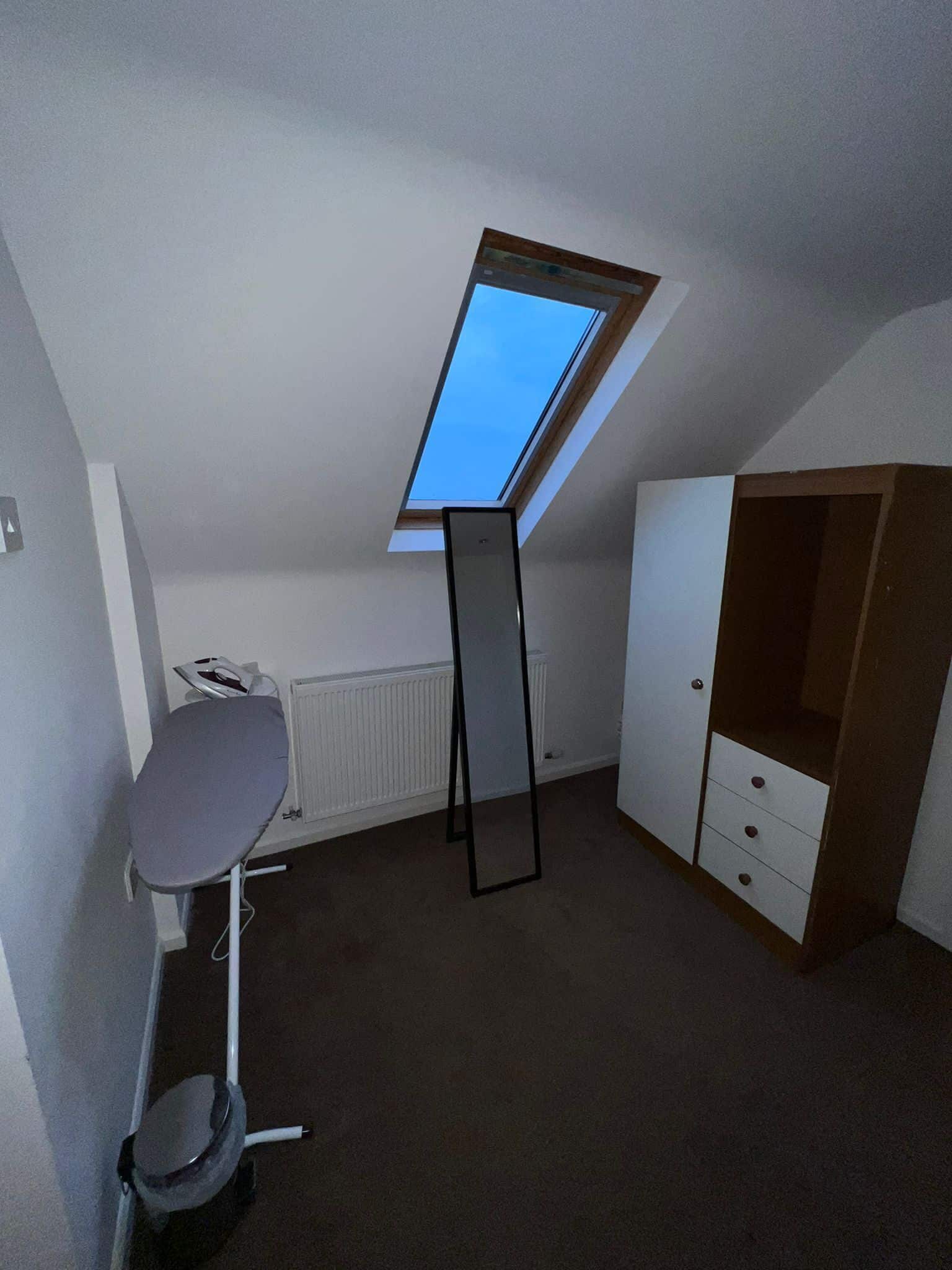 A room with a slanted ceiling, a radiator, a desk with a chair, a wardrobe, and a small trash bin.