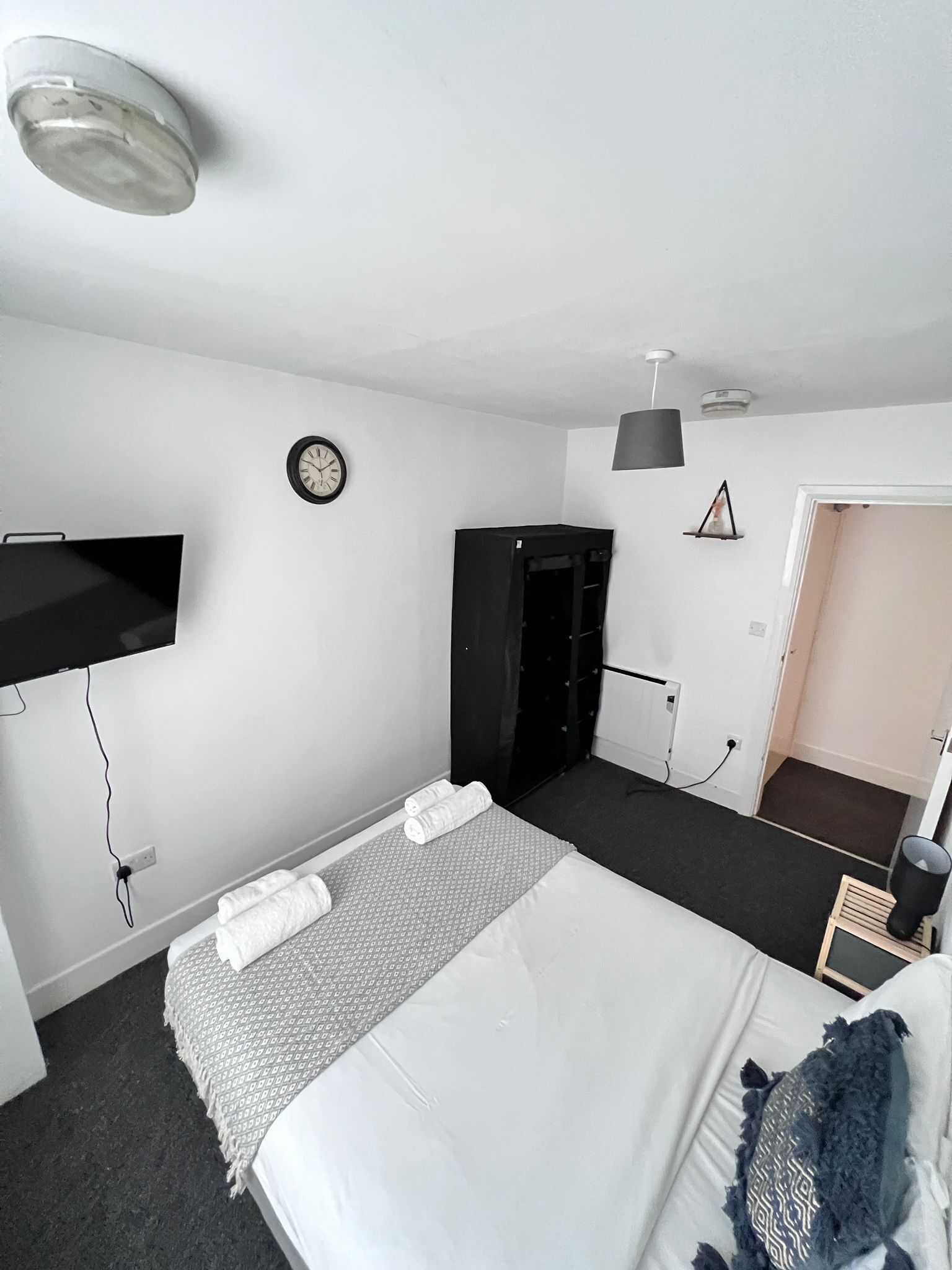 A hotel room with a bed, a wardrobe, a TV mounted on the wall, and a clock.