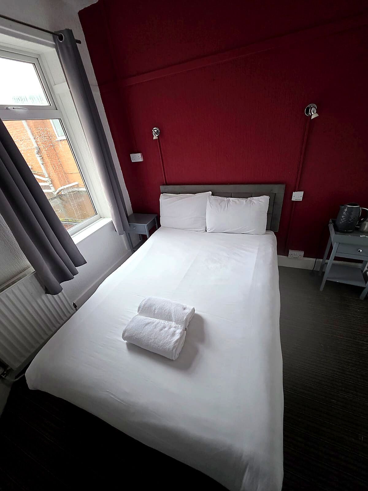 A hotel room with a large bed, white linens, a red accent wall, a window with curtains, and a small table with a kettle.