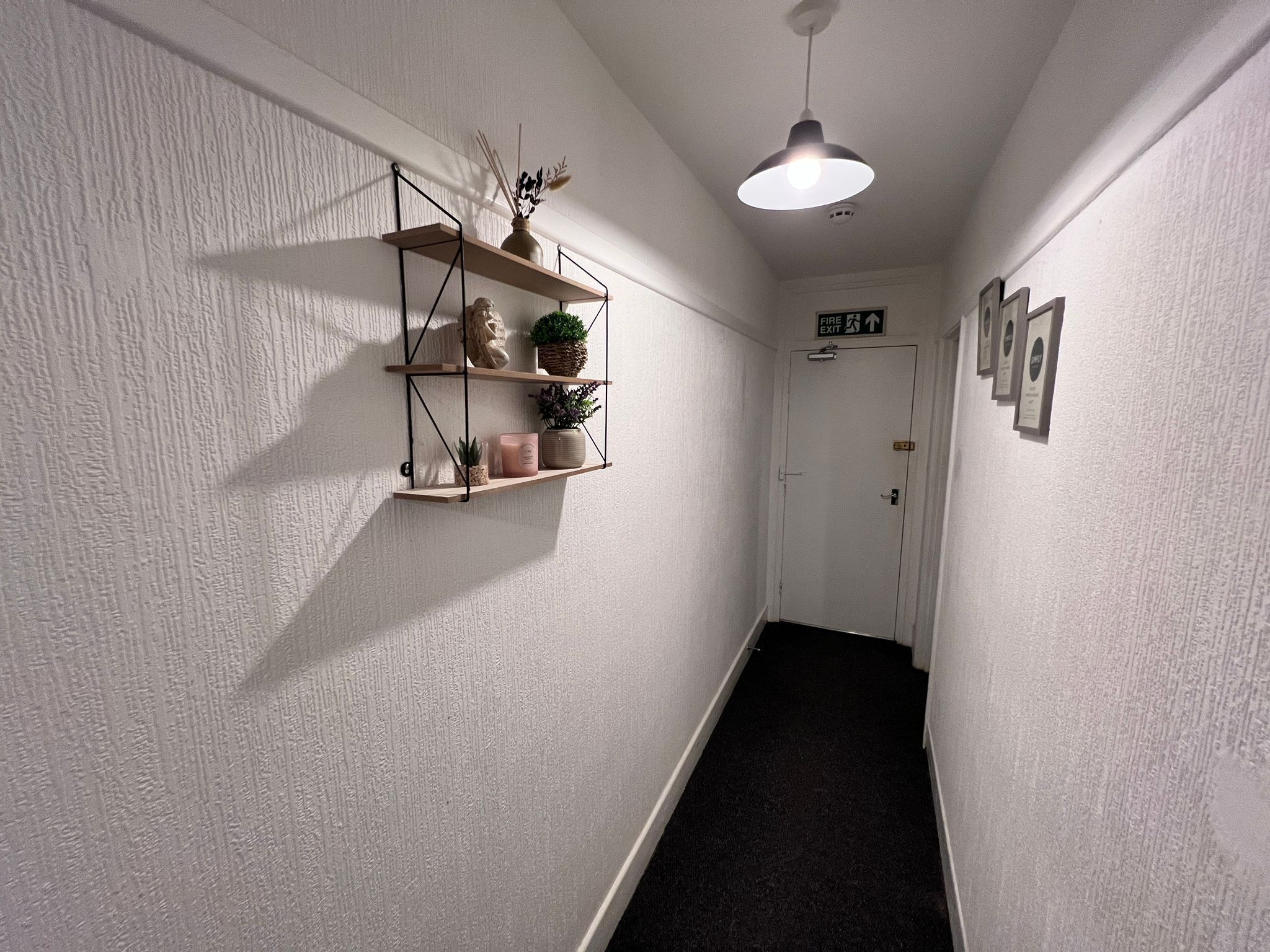 A hallway with a white textured wall, a wooden shelf with plants and candles, a white door with a fire exit sign, and a pendant light.