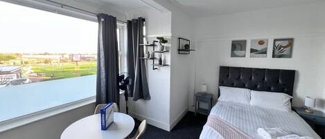 A hotel room with a bed, a desk, a chair, a small table, a nightstand, a lamp, a fan, a window with curtains, and a view of a golf course.