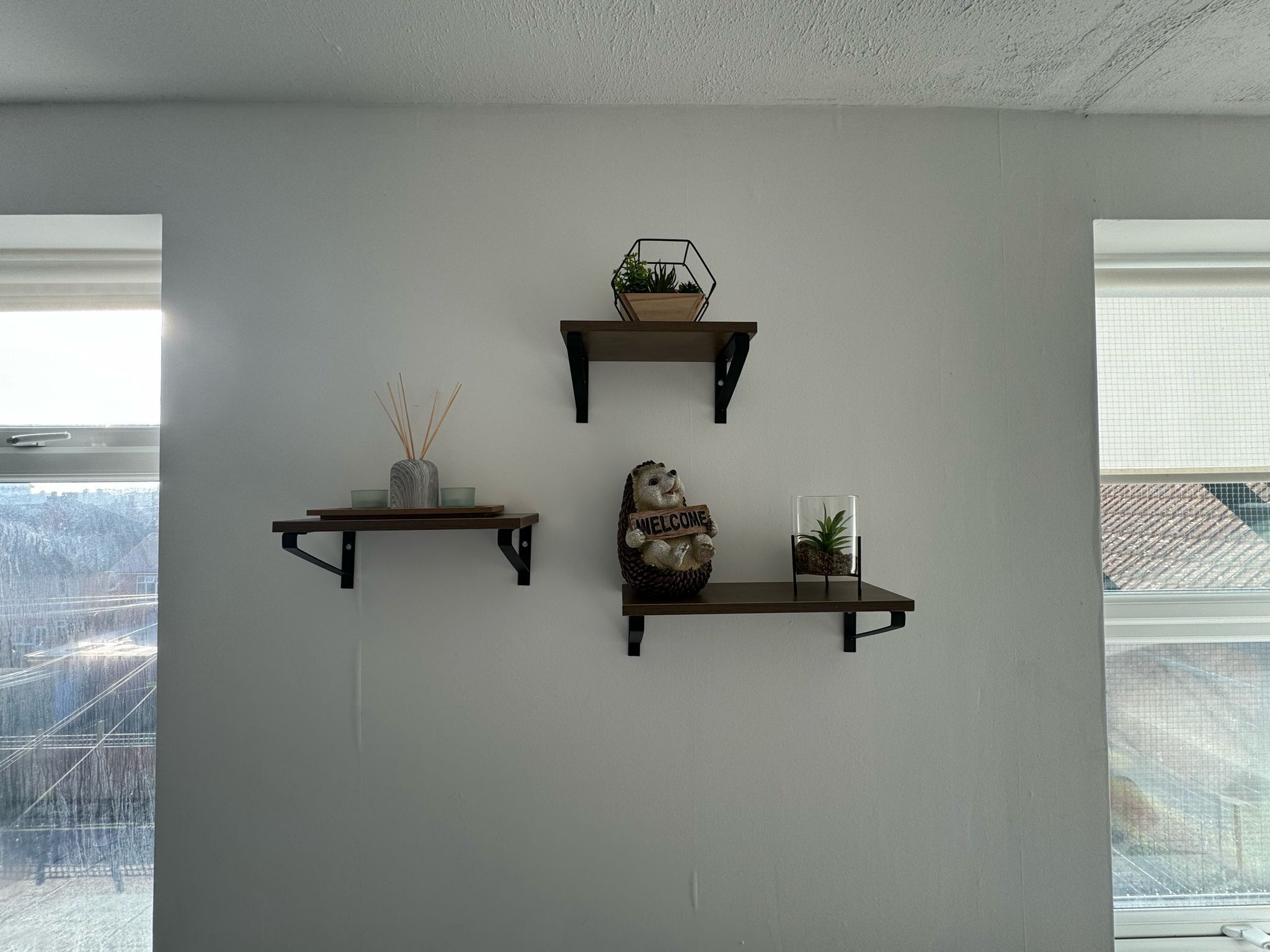 Três prateleiras fixadas na parede com diversos objetos decorativos, incluindo uma placa de boas-vindas, uma pequena planta e um vaso com juncos.