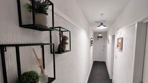 A hallway with white textured walls, black-framed mirrors, and a dark carpet. There are potted plants and a hanging light fixture. - Harbour Stays (Skegness)