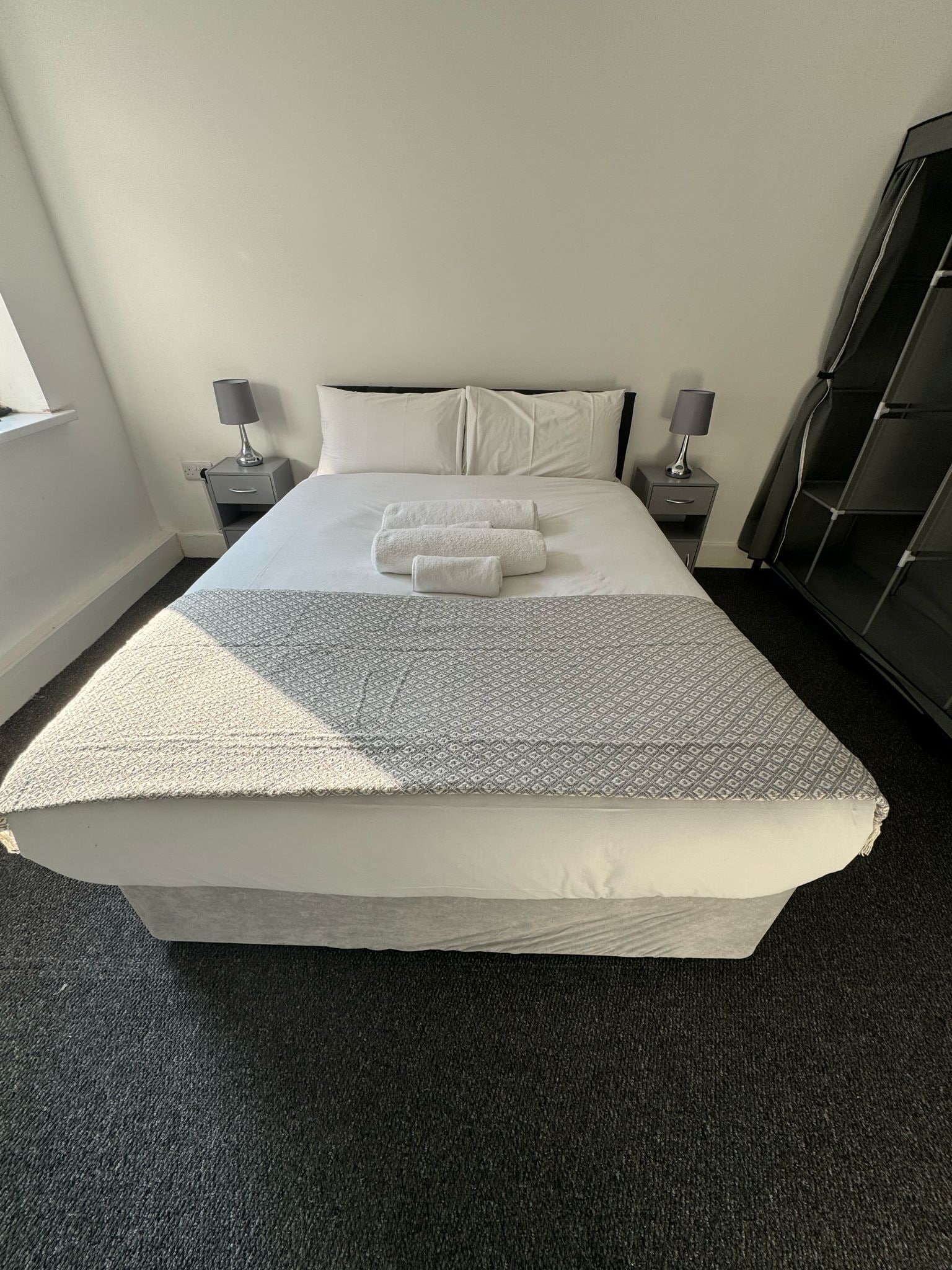 Una cama bien hecha con una colcha estampada y dos toallas dobladas. Hay dos mesitas de noche con lámparas, un armario de color oscuro y una ventana con marco blanco.