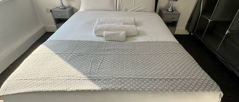 Una cama bien hecha con una colcha estampada y dos toallas dobladas. Hay dos mesitas de noche con lámparas, un armario de color oscuro y una ventana con marco blanco.