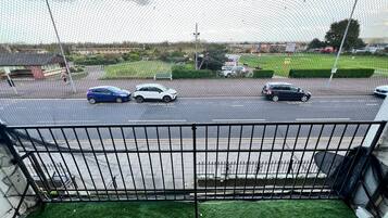 Vista desde un balcón con baranda de metal, que da a un estacionamiento con varios coches, un campo verde y un edificio con un letrero que dice "PUTTING COUPONS".