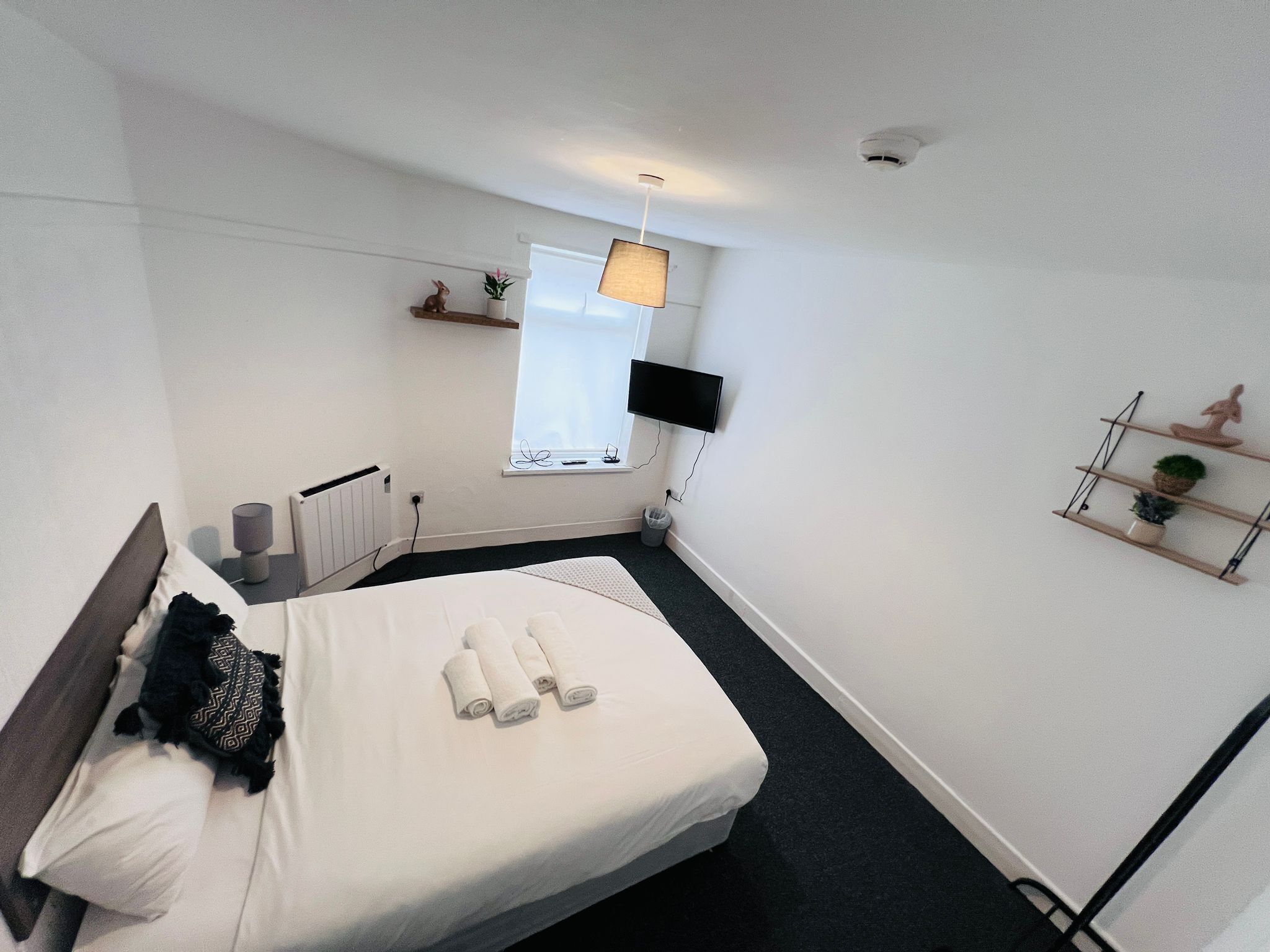 A modern hotel room with a bed, bedside table, lamp, window, and wall-mounted TV.