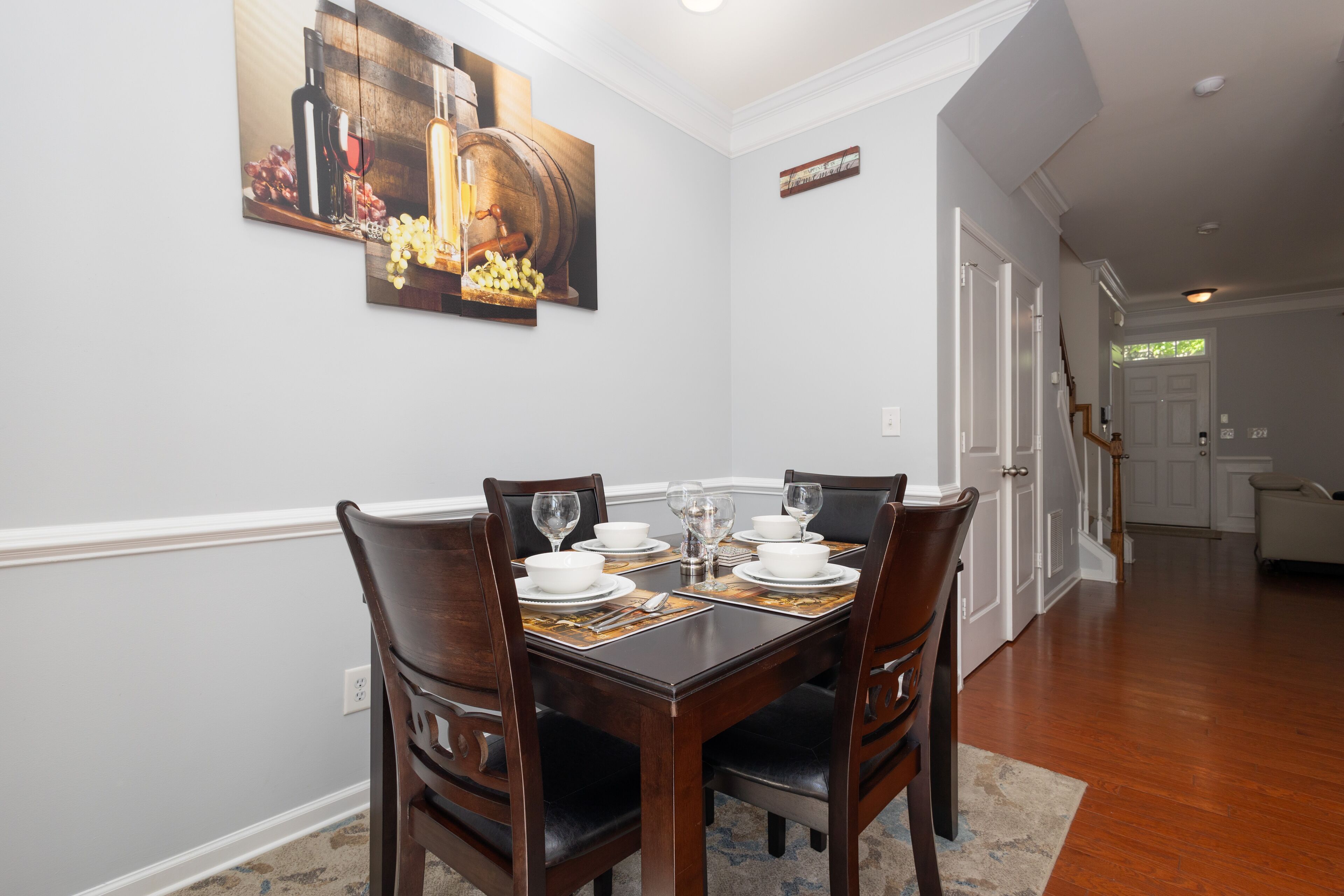 Une salle à manger avec une table en bois dressée pour quatre personnes, des murs blancs et un tableau représentant des bouteilles de vin et des grappes de raisin.