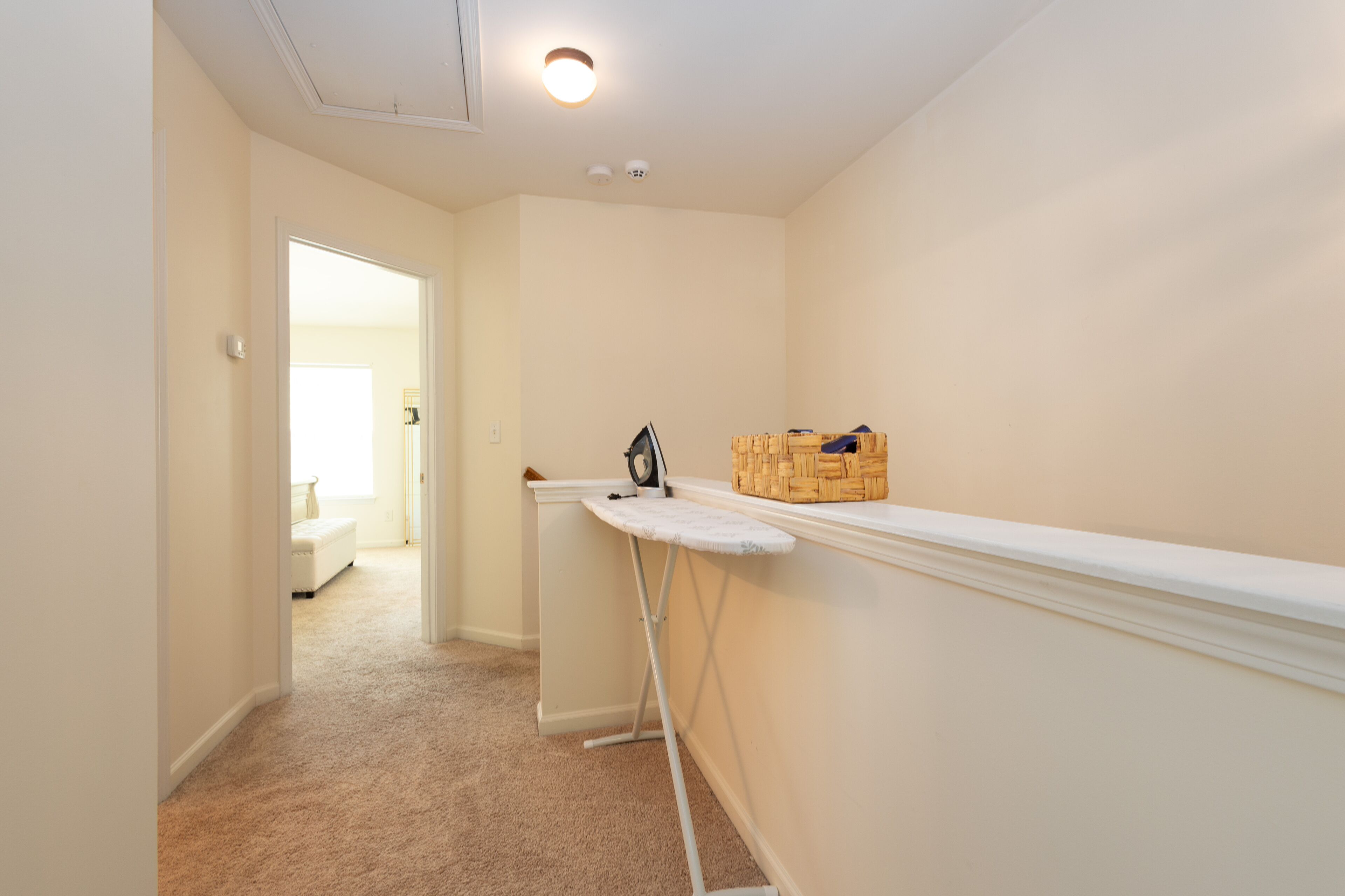 A hallway with a white ironing board, a basket of folded clothes, and a door leading to another room.