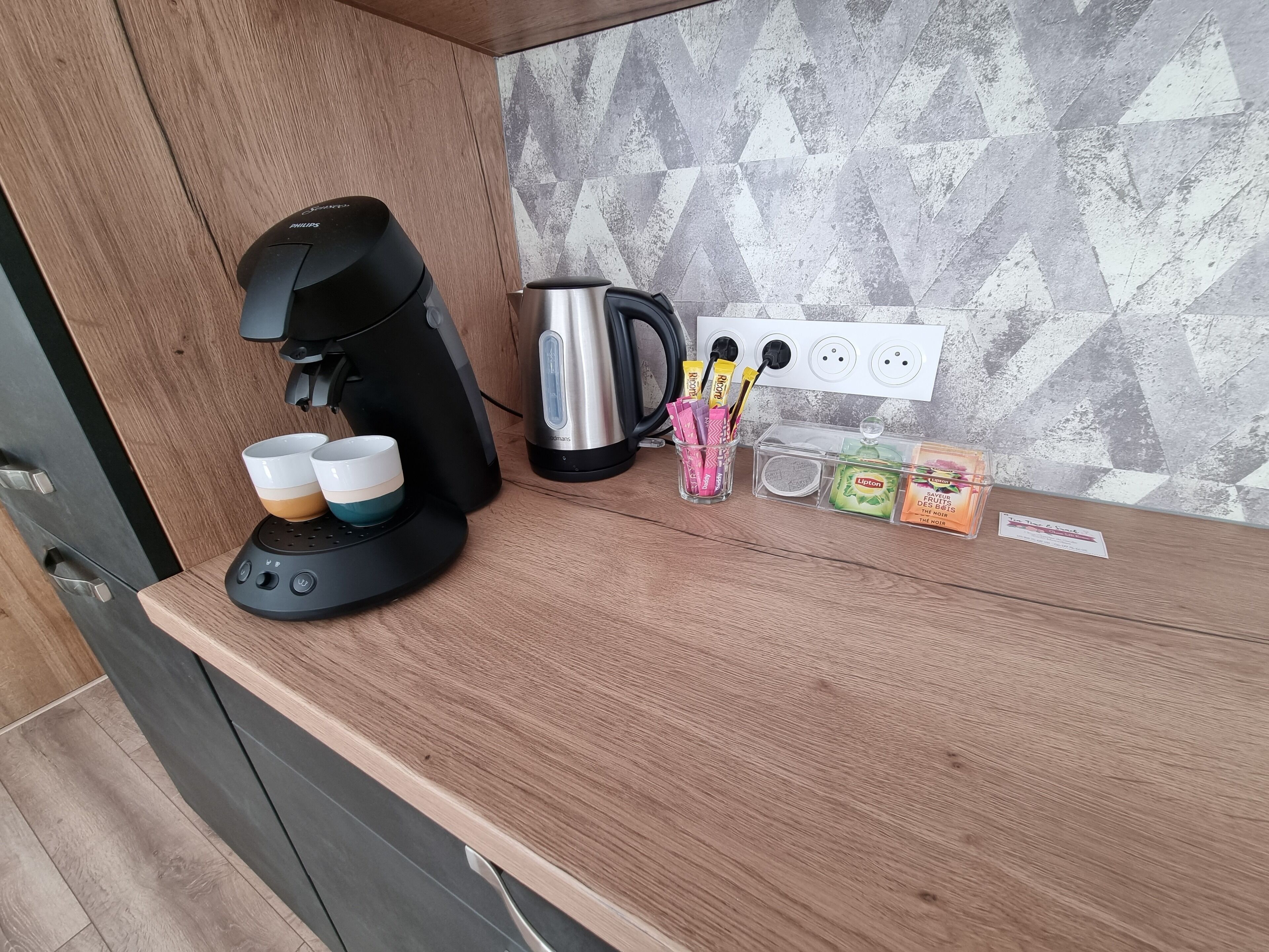 Eine Küchenzeile mit Kaffeemaschine, Wasserkocher und einer Tassehalterung inklusive Tassen. Außerdem befinden sich Gewürze und eine kleine Schachtel auf der Arbeitsfläche.
