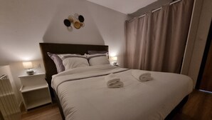 A neatly made bed with white linens and two towels folded at the foot. There is a nightstand with a lamp and a small table beside the bed. The room has a wooden floor and a curtain on the window. - Résidence Cosy et Moderne (Morvillars)
