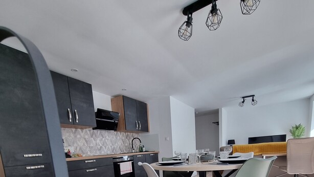 A modern kitchen with dark cabinets, a central island, and a dining area with a table and chairs. - Résidence Cosy et Moderne (Morvillars)