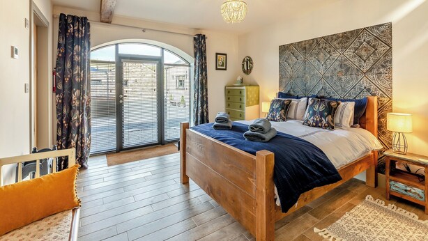 Ein Schlafzimmer mit einem hölzernen Bett, einem gemusterten Kopfteil, einer Chandelüster und Blick durch eine Glastür.